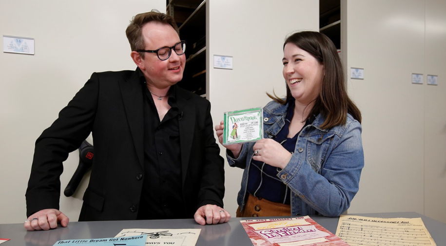 Professor Dominic Broomfield-McHugh in a black v-neck and sports coat smiles as he looks down at a CD of Donnybrook held by Director of Library and Archives Emily Rapoza in a jean jacket, patterned blue shirt and tan skirt.